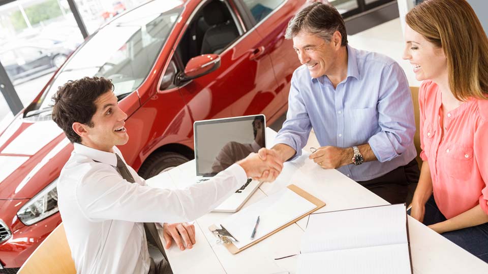 Ein Autoverkäufer schüttelt den Kunden die Hand in einem Autohaus in Baden-Baden, im Hintergrund ist ein rotes Auto zu sehen und auf dem Tisch liegen Unterlagen.